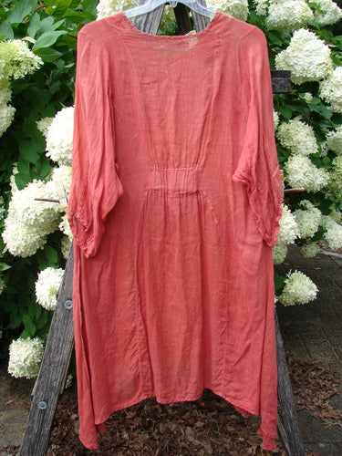 Bluefishfinder.com Barclay Linen High Vent Venetian Dress Center Starburst Poppy Size 1 in a muted poppy-red linen, shown as a loose long-sleeve dress with a center seam, gathered waist, and high vent details. The soft, lightly wrinkled fabric hangs from a hanger, framed by white hydrangeas and dark wood.
