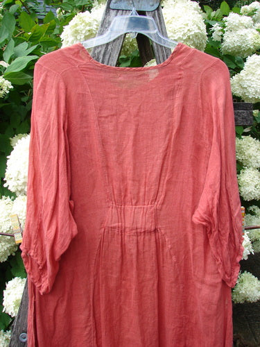 Bluefishfinder.com Barclay Linen High Vent Venetian Dress Center Starburst Poppy Size 1 in a soft poppy-red linen, hanging on a clear plastic hanger. The dress has a round neckline, airy semi-sheer texture, gathered waist, and loose three-quarter sleeves against white hydrangeas and weathered wood.