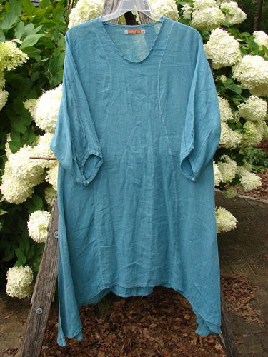 Bluefishfinder.com Barclay Linen High Vent Venetian Dress in unpainted jade, a loose linen garment with a V-notch neckline, three-quarter sleeves, and high side vents. The softly wrinkled fabric hangs from a hanger, outlined against dense green foliage and white blooms.