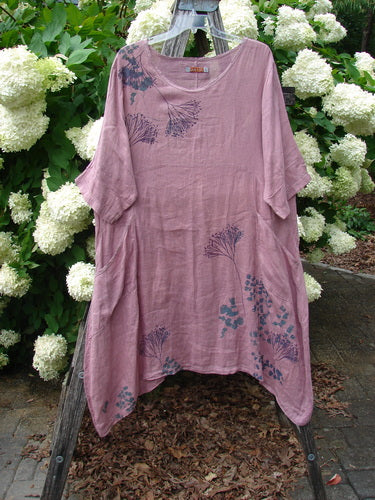 Bluefishfinder.com Barclay Linen Wave Pocket Dress in Dried Sprig Tulsi Rose, Size 1, shown as a loose linen dress with a soft rose-pink drape, short sleeves, and an airy, asymmetrical hem. Dark botanical prints scatter across the front and lower skirt as it hangs from a wooden stand amid white hydrangeas and green foliage.