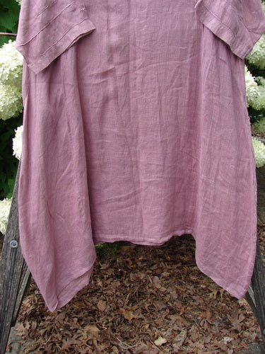 Bluefishfinder.com Barclay Linen Wave Pocket Dress in Dried Sprig Tulsi Rose, size 1, a loose mauve linen dress with a softly crinkled texture, short sleeves, and a rounded hem. It hangs centered against an outdoor backdrop, with muted green foliage and wood framing the lower edge.