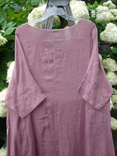 Bluefishfinder.com Barclay Linen Wave Pocket Dress in dried sprig tulsi rose, shown as a loose linen dress with a soft, wrinkled texture, wide three-quarter sleeves, and a simple rounded neckline. The muted rose fabric hangs from a clear hanger, with hydrangea blooms and greenery behind it.