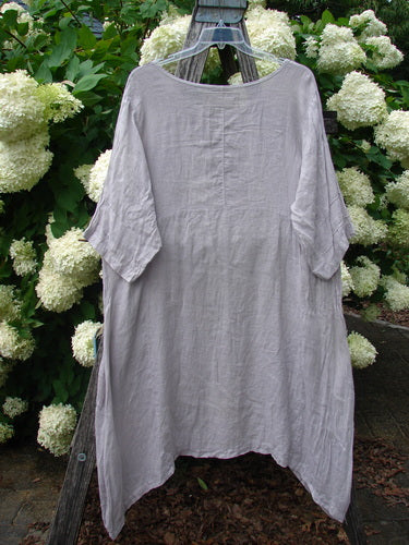Bluefishfinder.com Barclay Linen Wave Pocket Dress Fern Rice Paper Size 1 in soft rice-paper gray linen, with a loose, airy silhouette, short sleeves, and a gently uneven hem. It hangs centered on a wooden ladder against white hydrangeas and green foliage.