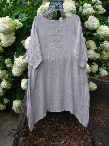 Bluefishfinder.com Barclay Linen Wave Pocket Dress in fern rice paper, size 1, hangs on a wooden stand. The loose, asymmetrical linen dress has a soft gray-lavender tone, crinkled texture, short wide sleeves, and a relaxed drape against white hydrangeas and green foliage.