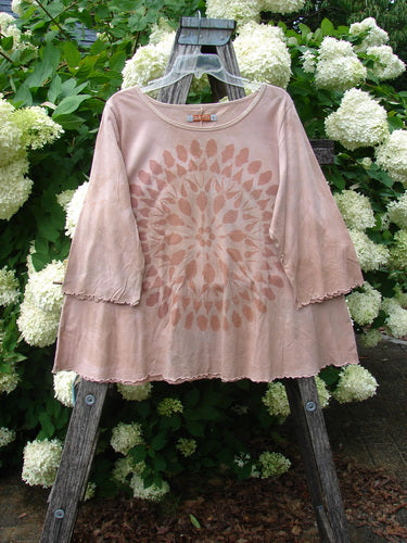 Bluefishfinder.com Barclay Three Quarter Sleeved Cafe Tee Giant Sunflower Mottled Pumpkin Size 1 in a soft mottled pumpkin tone, with three-quarter sleeves, raw-edged hems, and a large sunflower motif centered on the front. The lightweight tee hangs from a wooden easel amid dense white hydrangeas, its muted rose-beige fabric and airy drape standing out against the lush green backdrop.