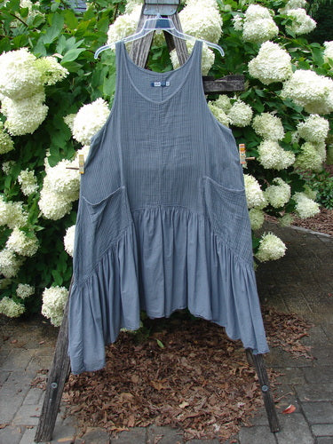 Bluefishfinder.com Barclay Gauze Batiste Summer Frock Jumper in unpainted gray day stripe, a sleeveless dress with a soft gauze-batiste texture, gathered skirt panels, and a loose, airy drape. It hangs on a wooden stand outdoors, framed by white hydrangeas and green foliage under muted natural light.