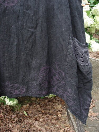 Bluefishfinder.com Barclay Summercloth Lace Two Pocket Pinafore in Moody Night Bloom Black, size 0, shown in a dark, textured fabric with lace-trimmed pocket detail and a faint floral bloom near the hem. The skirt falls in a soft, uneven drape, with the black and muted purple pattern standing out against the outdoor background.