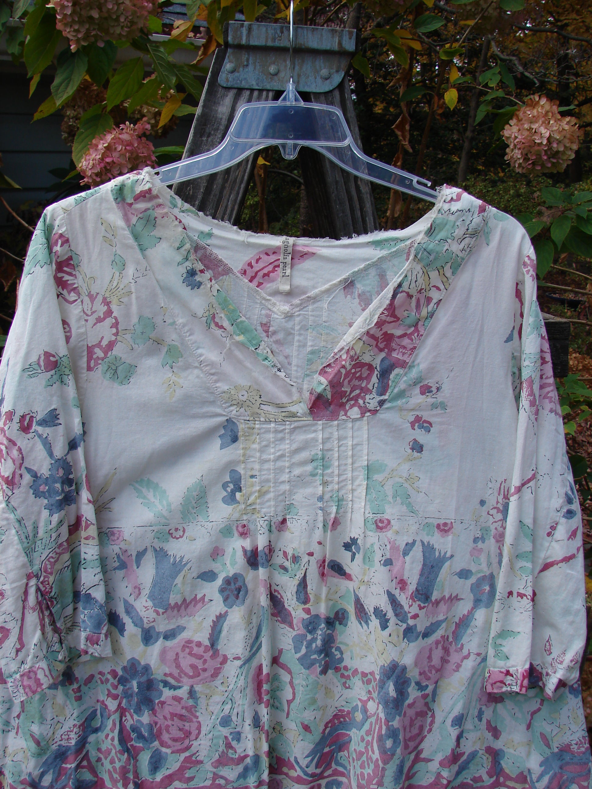 Magnolia Pearl European Cotton Breeze Blouse Spring Florals OSFA, featuring a floral pattern on lightweight linen-cotton, displayed on a hanger, showcasing pleats, raw edges, and an A-line shape.