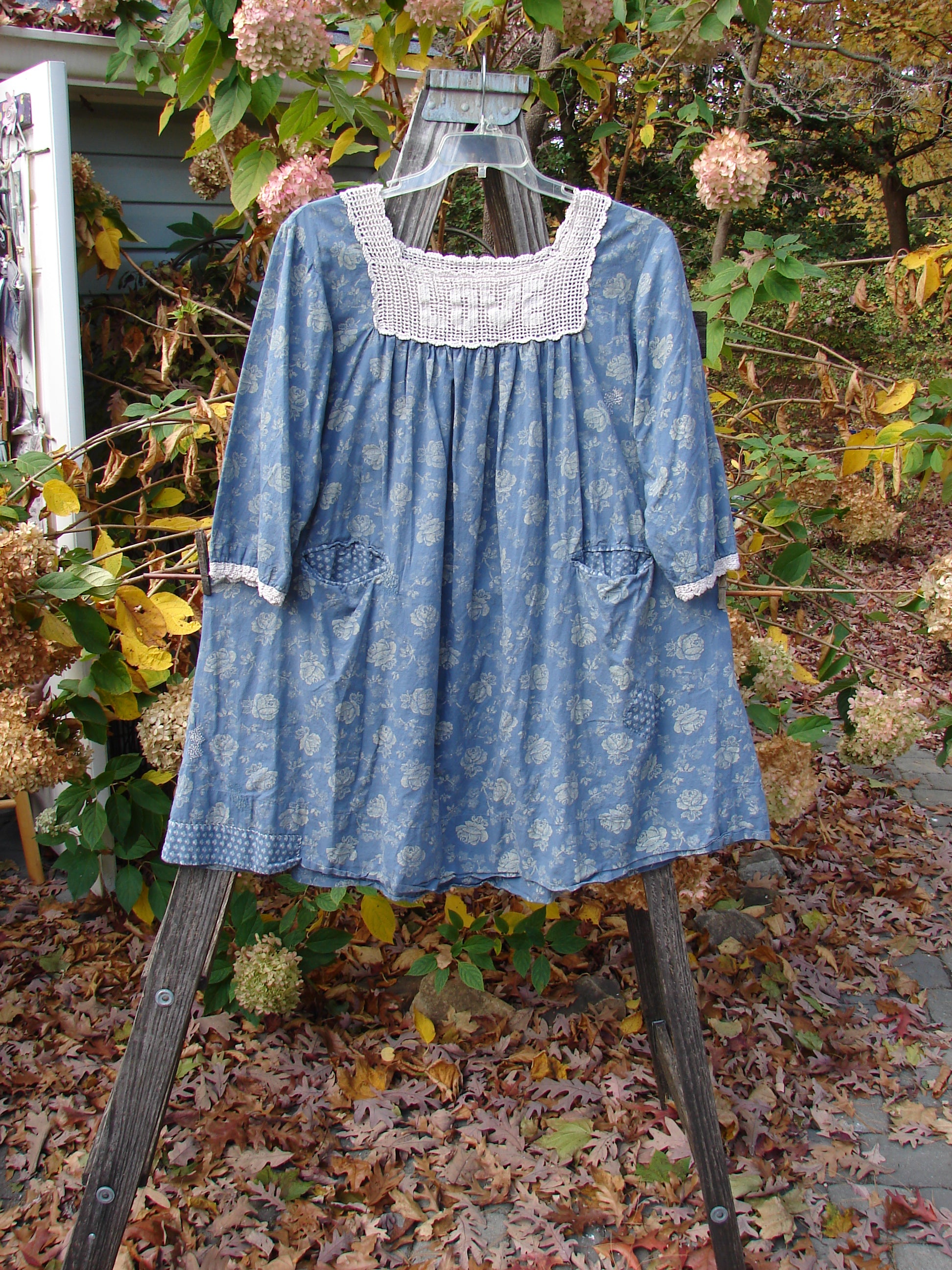 Magnolia Pearl European Cotton Crochet Love Tunic Vintage Blue Rose OSFA displayed on a swing, showcasing its gathered crochet top, three-quarter sleeves, and floral interior pockets against a leafy backdrop.