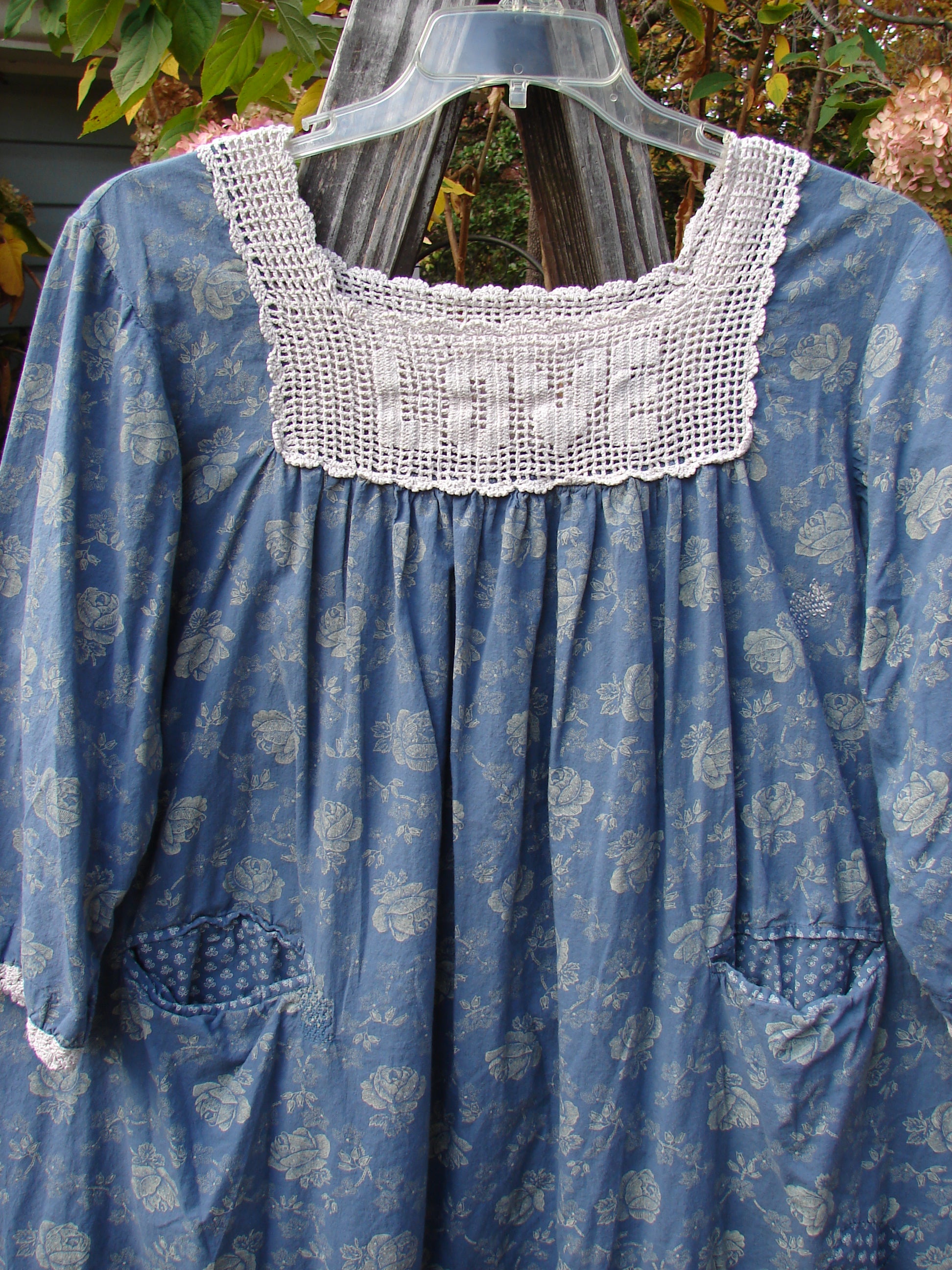 Magnolia Pearl European Cotton Crochet Love Tunic Vintage Blue Rose OSFA displayed on a hanger, showcasing its intricate white crochet lace, three-quarter sleeves, and A-line design against a vintage blue rose backdrop.