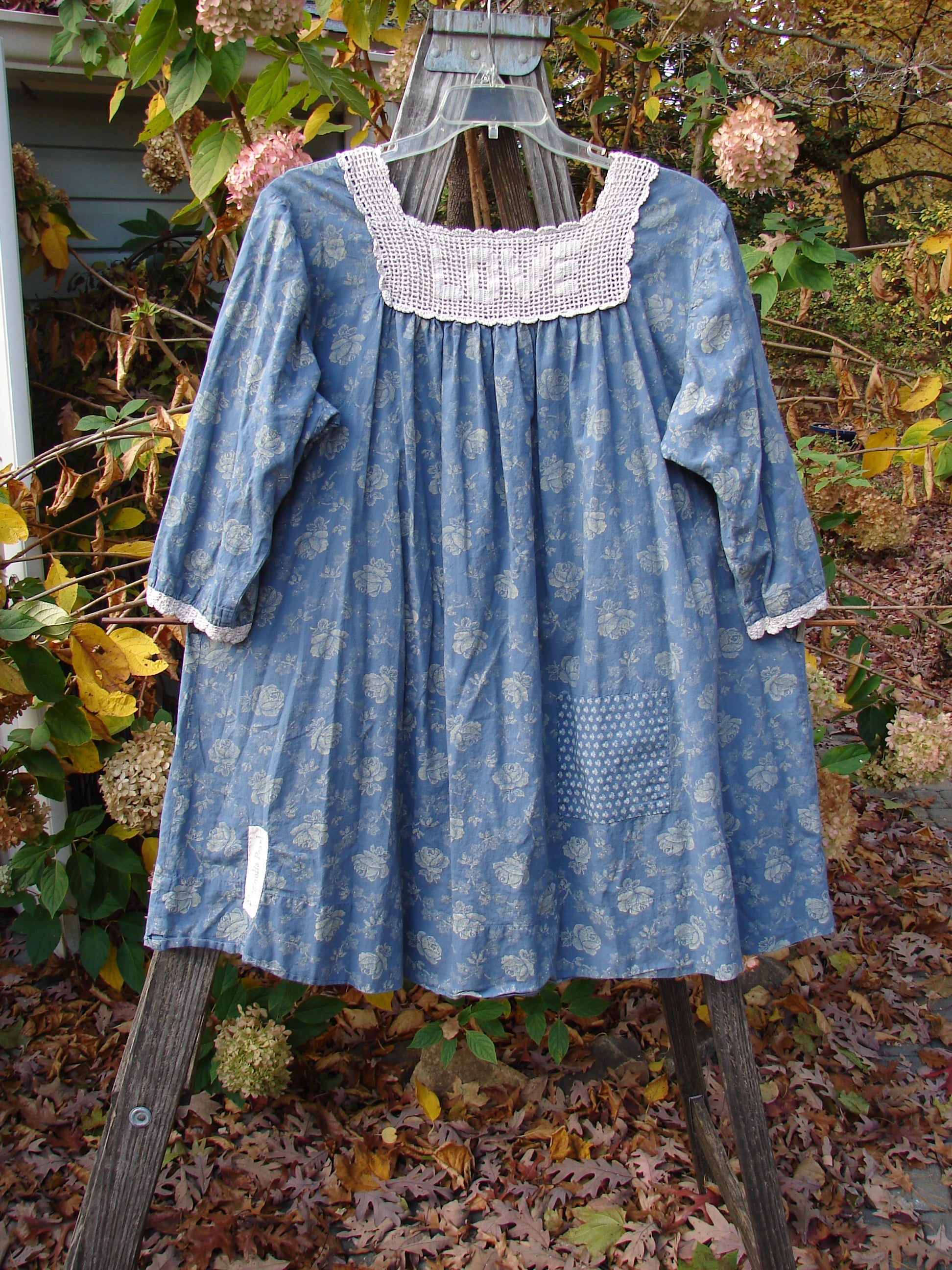 Magnolia Pearl European Cotton Crochet Love Tunic Vintage Blue Rose OSFA displayed on a swing, showcasing its crochet square top, floral interior pockets, and A-line shape with a vintage blue rose pattern.