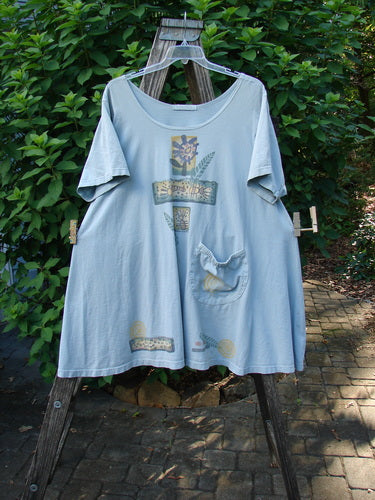 1995 Hop Scotch Dress Garden Friends Beach Glass Size 2 displayed on a wooden stand, features a light-blue oversized T-shirt design with decorative patches, wide scooped neckline, and elastic-topped pocket.
