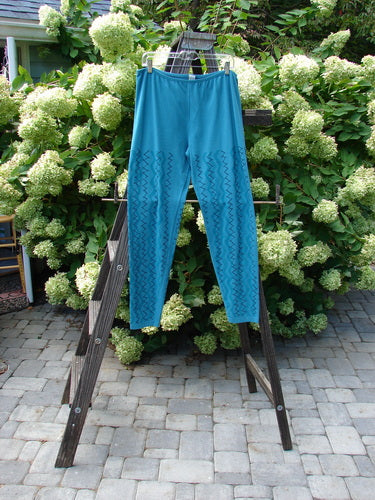 Barclay Cotton Lycra Bally Layering Pant Legging in Aqua, size 2, on a ladder in a garden. Full elastic waistband, slightly narrowing lower, average length legging. Continuous criss-cross theme paint. Soft forgiving feel, Blue Fish patch.