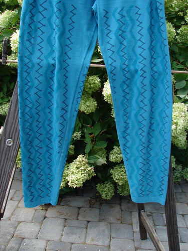 A Barclay Cotton Lycra Bally Layering Pant Legging in Aqua, featuring a pair of blue pants on a rack. Perfect condition, medium weight fabric with a full elastic waistband, slightly narrowing lower, and average length legging. A fun layering piece with a continuous criss-cross theme paint. Soft and forgiving feel, with the interior signature Blue Fish patch. Waist relaxed 28, fully extended 40, hips 42, inseam 29, and length 40 inches.