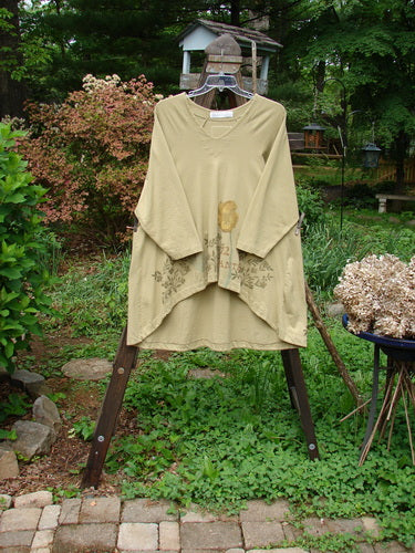 1998 Botanicals Bell Flower Top in Stem on a rack, featuring a long-sleeved beige shirt. Close-ups of a plant, metal pole, bush, and sidewalk add visual interest.