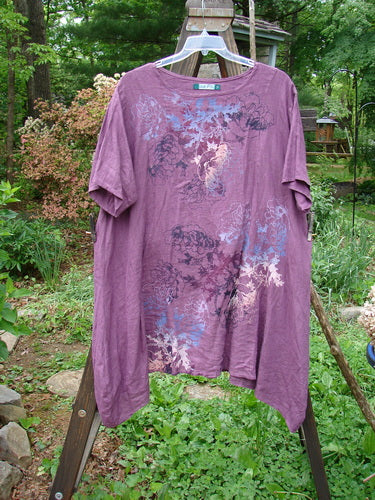 Barclay Linen Figure 8 Tunic Dress Garden Shadow Red Plum Size 2: A purple shirt on a swinger with a floral design, paired with a close-up of a green plant.