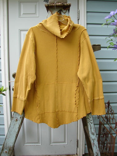 A yellow sweater on a ladder, featuring a curly cowl neck and a kangaroo tunnel pocket. The Barclay Thermal Reverse Stitch Tunnel Pocket Top in unpainted mustard is a unique and cozy addition to your wardrobe.