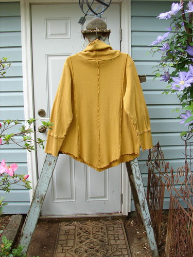A yellow jacket on a ladder, wearing the Barclay Thermal Reverse Stitch Tunnel Pocket Top in Mustard. The top features a curly edged floppy turtleneck, a kangaroo tunnel pocket, and stitched vertical sectional seams. It has a unique cut with changes in just the right places.