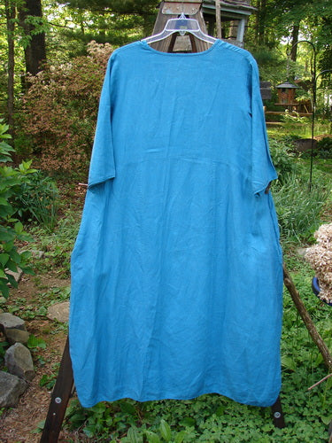 A Barclay Linen Long Urchin Dress in Dusty Aqua, featuring a rounded neckline, downward curved empire waist seam, and generous three-quarter sleeves. The dress has an overall widening shape, deep side pockets, and a unique continuous wave theme paint. Size 1.