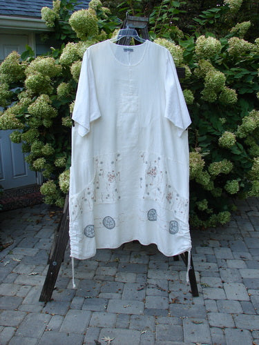 A white Barclay Linen Farmer Jen Dress with a floral design on the sleeves. Features include a sectional bodice, wrap around pockets, and a paneled lower hem. Size 2.