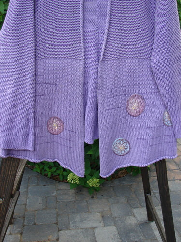 1999 Rollneck Cardigan Sweater: A purple sweater with flowers, dual knitting, rolled seams, and a varying hemline. Features a cropped back line and porcelain button closure.