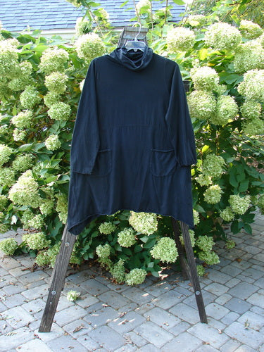 A black tunic top with a cowl neck, double pockets, and curly edges. Made from organic cotton, it features empire waist seams, lettuce edgings, and a varying hemline. Size 0.