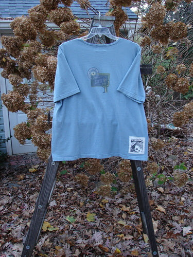 2000 Short Sleeved Tee Greetings from Santa Fe Blue Jet Size 1: A blue shirt with a logo on it, standing on a wooden stand outdoors.