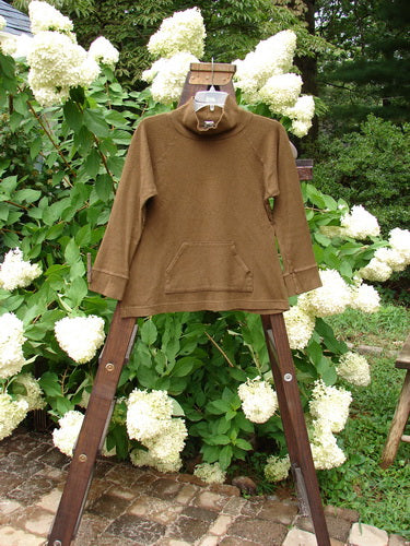 2000 Wool Funnel Neck Raglan Top Unpainted Pebble Smaller Size 1: A brown sweater on a wooden stand, featuring a relaxed half-height turtleneck, banded lower cuffs and hemline, and a front center cargo pocket.