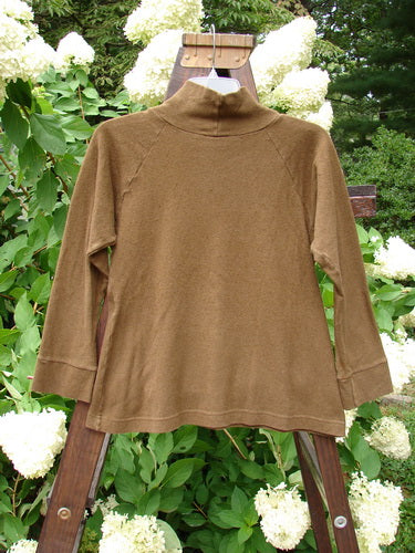2000 Wool Funnel Neck Raglan Top Unpainted Pebble Smaller Size 1: A brown sweater on a wooden stand, featuring a relaxed half-height turtleneck and a front center cargo pocket.