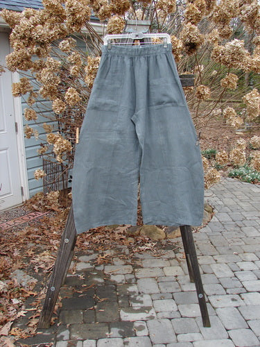 A pair of Barclay Linen Crop Side Pocket Pants in Dusty Sky, size 0, on a wooden stand. Features include a thicker elastic waistline, billowing fall, and upper hip front pockets. Unpainted for easy mixing and matching.