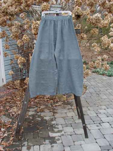 A pair of Barclay Linen Crop Side Pocket Pants in Dusty Sky, size 0, on a wooden stand. Features include a thicker elastic waistline, billowing fall, sectional panels, and upper hip front pockets. Unpainted for easy mixing and matching with other pieces.