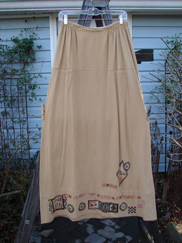Image alt text: 1998 Scrabble Skirt Games Bamboo Size 2 - A long beige skirt with a cartoon on it, featuring a waistline with folded front and rear elastic, horizontal panels, a lower banded wider sweep, and two upper finger pockets.