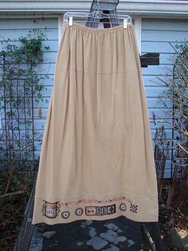 1998 Scrabble Skirt Games Bamboo Size 2: A long beige skirt on a clothes rack with a waistline, horizontal panels, and a lower banded wider sweep. Features classical games theme paint and a serious lower weighted swing.