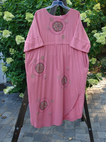 Barclay Naiad Dress Medallion Glow Size 2: A pink dress with a pattern, featuring a tapered lower shape, generous bust, rear draw cord, shorter sleeves, and a rounded, vented hemline. Includes deep side pockets and the Barclay Medallion Theme Paint. Made from mid-weight organic cotton. Length: 54".