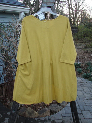 A yellow high low top on a clothes rack, featuring a rounded banded bottom shape, feminine neckline, and varying hemline for a full bottom swing. Size 2, unpainted.