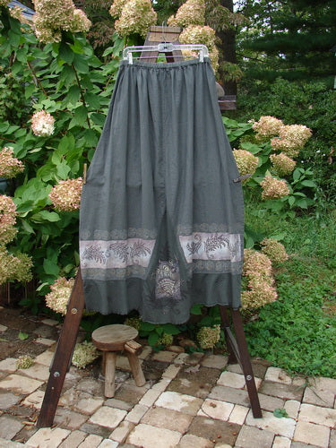 Barclay Linen Duet Skirt Green Raven Size 2: A pair of pants and a long grey skirt with a floral design on a rack, with a wooden stool on the ground.