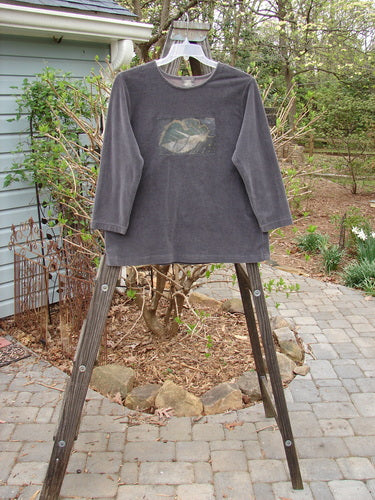 1999 Patched Stretch Cord Pullover Top Leaf Grey Plum Size 0: A long-sleeved shirt on a rack with a wooden ladder. The shirt features a painted front patch in a leaf theme. It has a rounded neckline, drop shoulders, and a slight A-line shape. Made from stretch cotton corduroy, this cozy piece is perfect for fall.