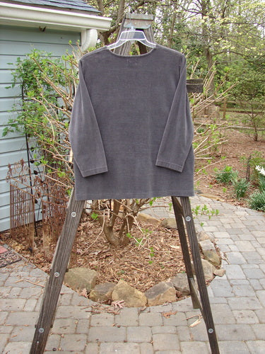 1999 Patched Stretch Cord Pullover Top Leaf Grey Plum Size 0: A long-sleeved grey shirt on a wooden rack. Made from stretch cotton corduroy, it features a painted front patch with a single leaf theme. Cozy and comfortable, with a gentle rounded neckline and drop shoulders.