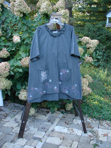 Barclay Linen Duet Sunrise Dress Tiny Rose Stormy Grey Size 2: A smock-style tunic with floral-embroidered pockets, gently ruffled neckline, and varying hemline, displayed on a wooden ladder in a garden setting.