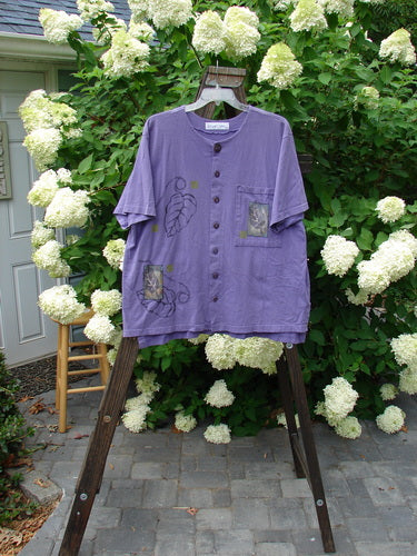 1998 Camp Shirt Falling Leaf Modena Size 2: A purple shirt with a leaf design. Features include a button front, vented hemline, and oversized pocket. Made from organic cotton.