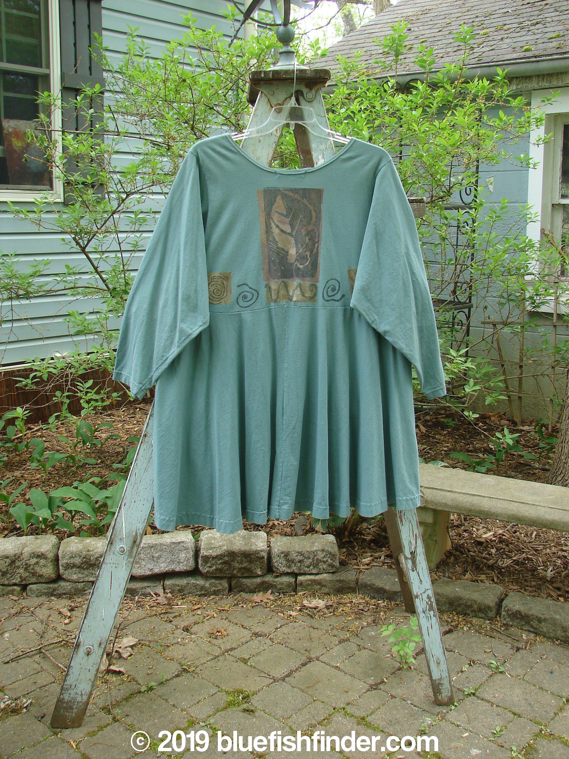 1994 Juniper Dress Swirl Leaf Sea Journey Size 2: A blue shirt on a wooden rack. Vintage cotton dress with rounded collar, recycled paper button closures, and a sweeping swing lower.