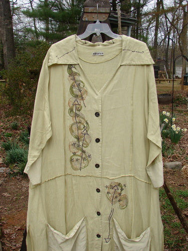 A long white dress with a design on it, featuring a deep V-shaped neckline, a prominent pointed collar, and contrasting fabrics of silk and linen. The dress has a varying vented hemline and unique super swirl buttons. It also has two silk contrasting and pinched drop hem pockets. The dress is painted in the Windy Star theme.