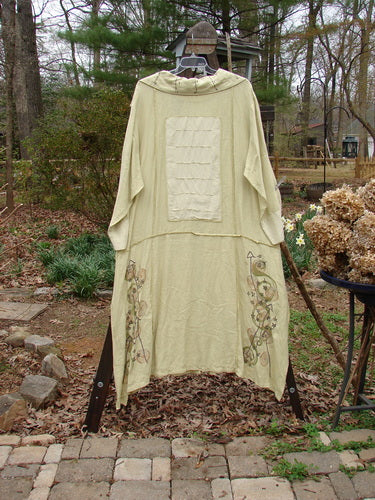 A long white robe with a white square on it, hanging on a rack. The robe is made from a mid-weight linen and silk combination. It features a deep V-shaped neckline, a prominent pointed collar, exterior horizontal stitchery and sectional panels, a varying vented hemline, contrasting fabrics of silk and linen, unique super swirl buttons, and two silk contrasting and pinched drop hem pockets. The robe is painted in the Windy Star theme.
