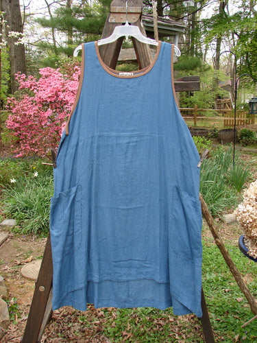Barclay Linen Contrast Pocket Jumper Unpainted Deep Teal Size 2: A blue apron on a wood stand, showcasing a super soft flowing shape with front wrap drop pockets and a sectional lower panel.