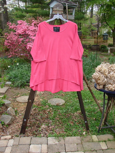 A pink Barclay High Low Top on a clothes rack, featuring a rounded banded bottom shape, feminine neckline, and varying hemline for a full bottom swing. Size 2, unpainted Flamingo.
