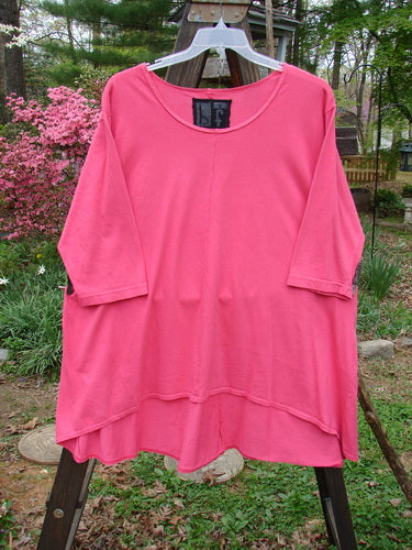 Barclay High Low Top Unpainted Flamingo Size 2: A pink shirt on a wooden stand, featuring a huge A-line rounded banded bottom shape, a thinner more feminine neckline, and a varying hemline for the best full bottom swing. The shirt has three-quarter inch sleeves and comes unpainted for easy mixing and matching.