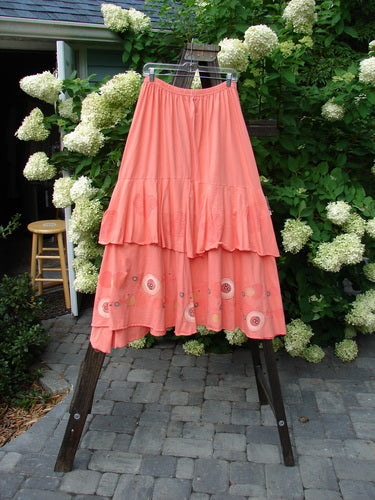 Image alt text: Barclay Two Story Skirt Bubbles Tangerine Size 2, featuring a pink skirt with sweet flutter-like edges on a mannequin, surrounded by plants and flowers on a stone patio.