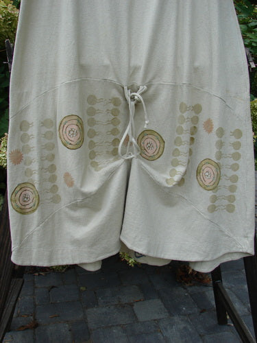 A close-up of a pair of 2000 Cotton Hemp Shade Skirt Bio Atom Dove shorts on a rack. The shorts have a full elastic waistband, an upward scoop and varying hemline when tied with the extra long rippie, and 3 vertical loops for manageable and creative adjustments. The shorts feature sectional horizontal panels and the classic Resort Biology Theme Paint. Waist Fully Relaxed 30 ~ Waist Fully Extended 52 ~ Hips 52 ~ Hem Circumference 70 and the Length is 37 Inches.