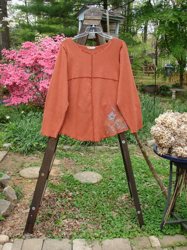 Barclay Cotton Lycra Curly Sectional Top Sea Shells Burnt Orange Size 2: Long-sleeved shirt on a wooden rack, with a close-up of a plant and a metal pole.