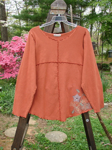Barclay Cotton Lycra Curly Sectional Top Sea Shells Burnt Orange Size 2: A long sleeved shirt on a rack, featuring a rounded widening hemline, drop shoulders, and double drop exterior pockets.